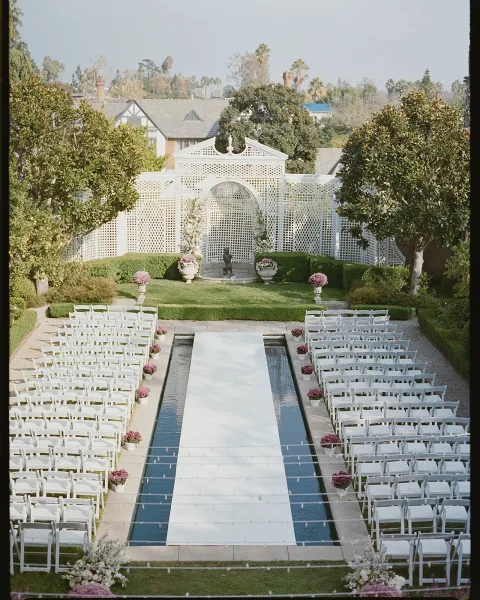 Ceremony setup for outdoor wedding ceremony with white aisle runner over a reflecting pool, lined by white folding chairs on a manicured lawn