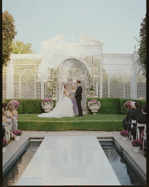 Ceremony moment as bride and groom hold hands at a poolside altar with white lattice backdrop and floral arch in a garden setting