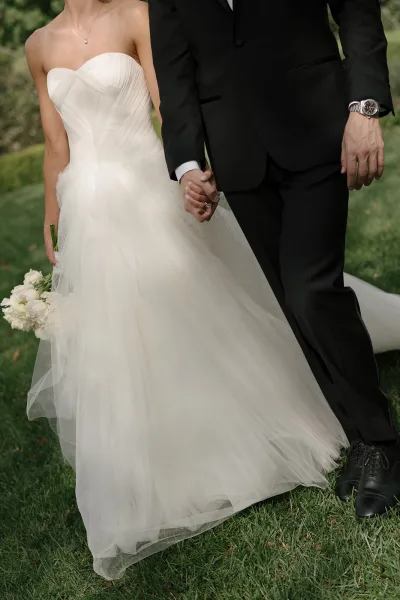 Couple portrait of bride and groom holding hands, showing wedding rings beside a white rose bouquet on a green lawn, tux and tulle dress visible