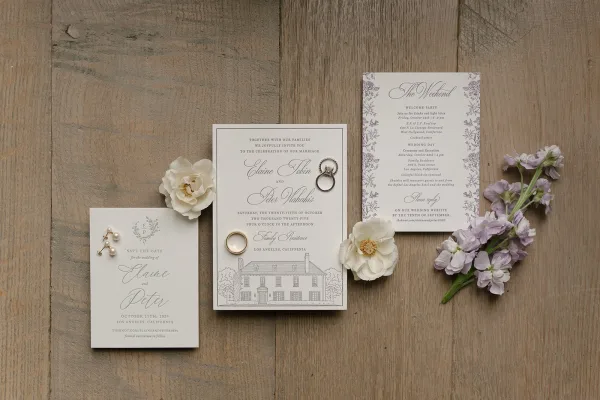 Invitation suite wedding invitation flat lay with save the date, rings, pearl earrings, and white and purple flowers on a rustic wood table