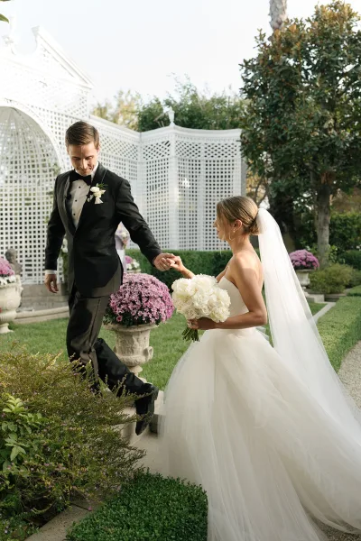 Couple portrait of bride and groom holding hands, bride with white bouquet and veil beside a white lattice gazebo on garden lawn
