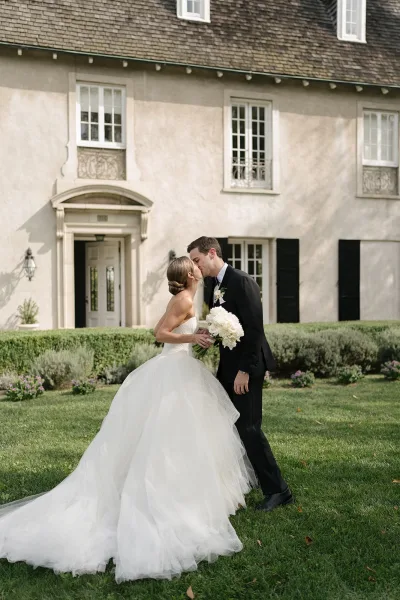 Wedding kiss as bride in a strapless tulle gown holds a white bouquet while groom in tux kisses her on a manicured lawn by an estate house
