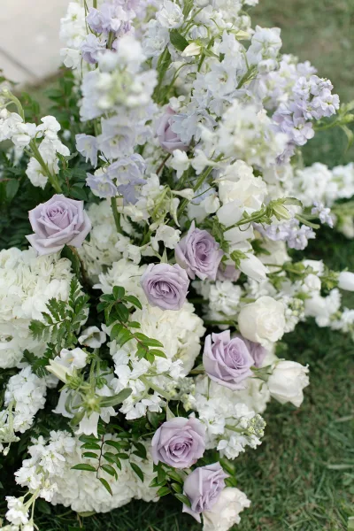 Wedding florals in lavender and white with roses, hydrangea, and lilac stock arranged low on a grass lawn for an outdoor aisle marker