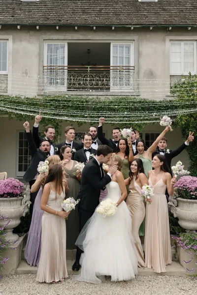 Wedding party photo of bride and groom kissing as friends cheer, bouquets raised, on ivy-covered courtyard steps with string lights overhead
