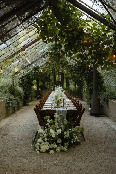 Reception tablescape with white rose centerpieces and greenery vines on a long wooden table, candles and string lights in a glass greenhouse