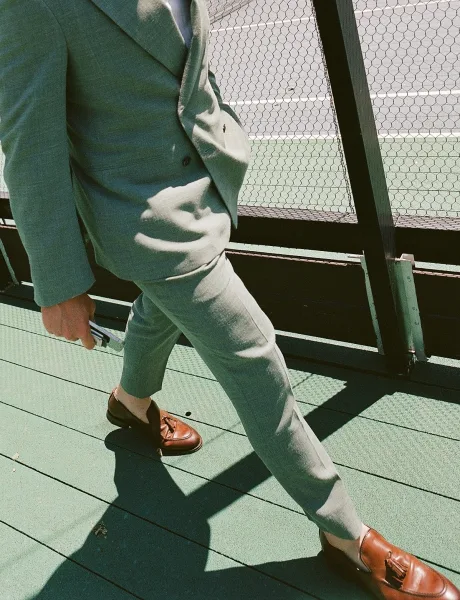 Groom suit captured in a candid walk, paired with loafers, along a painted sports court by a chain-link fence.