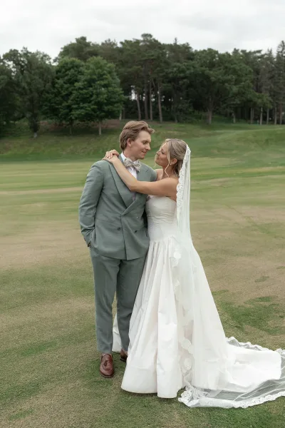 Couple portrait as the bride and groom embrace on a grassy lawn with trees under a cloudy sky, her lace veil softly draping behind them.