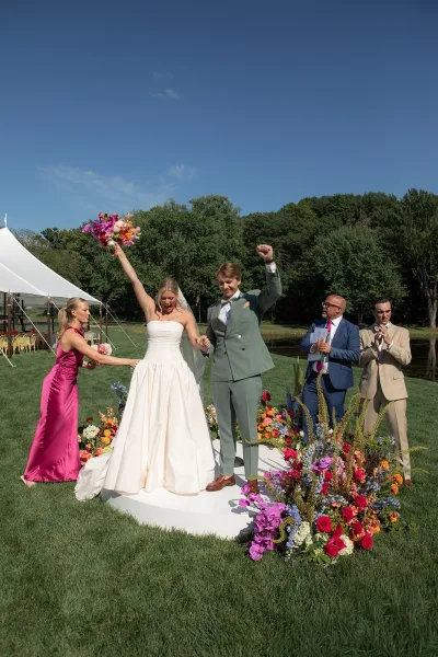 Ceremony moment during a just married celebration as the bride lifts her bridal bouquet and the groom fist-pumps beneath a sailcloth tent