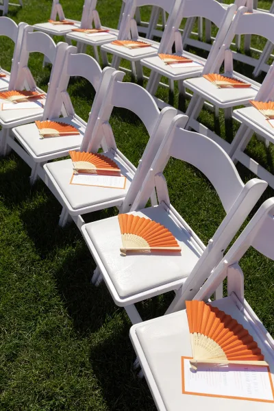 Ceremony seating for an outdoor ceremony setup arranged in neat rows, with hand fans laid on each chair across a simple grass lawn.