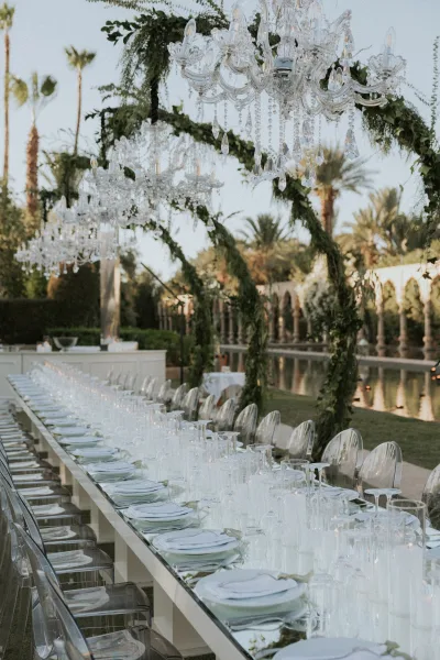 Reception tablescape with long banquet table wedding details, crystal chandeliers and hanging greenery by a reflecting pool with palm trees