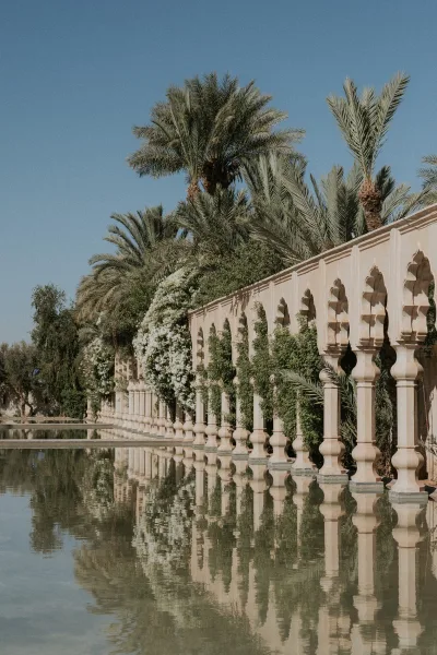 Wedding venue exterior with stone arches along a columned colonnade beside a reflecting pool, framed by palm trees and garden greenery