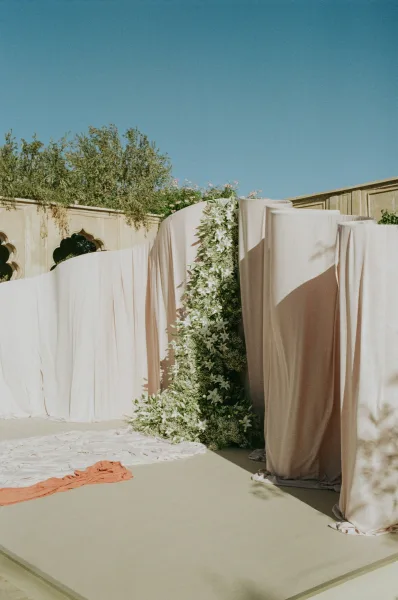 Ceremony backdrop with white floral installation and greenery accents on a stage, framed by ivory draped fabric in a sunny courtyard with arches