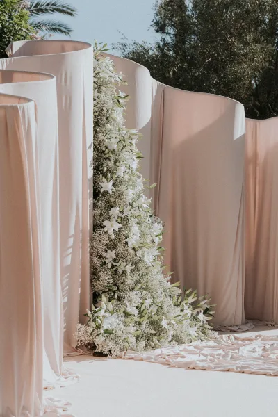 Wedding ceremony backdrop with draped fabric panels and white flowers, baby's breath, and greenery set against outdoor trees and sky