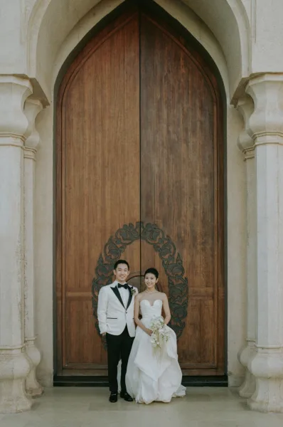 Couple portrait of bride in a strapless wedding dress and groom in a white tuxedo jacket holding a bouquet at arched wooden doors