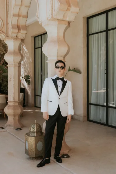Groom portrait in a white tuxedo jacket and black bow tie, wearing sunglasses on a stone-arched patio beside a decorative lantern