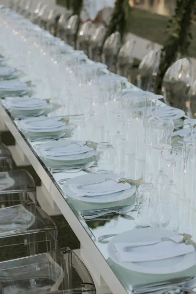 Wedding tablescape with white plates, folded napkins, silver flatware and clear glassware on a long table with transparent chairs on a lawn