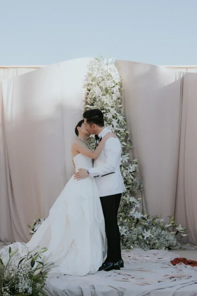Wedding kiss portrait of bride and groom kissing on an outdoor stage, framed by a floral backdrop of white lilies and baby's breath