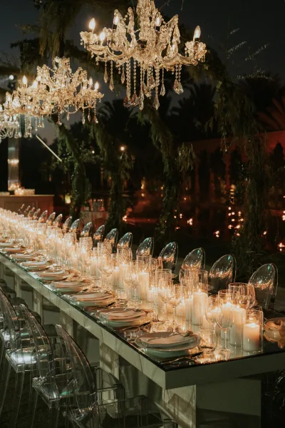 Reception tablescape with mirrored wedding table set for dinner, taper candles and glassware beneath crystal chandeliers and hanging greenery outdoors at night