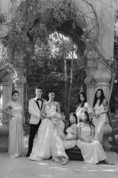 Wedding party portrait of bride and groom with bridesmaids under a vine-covered stone archway, bride holding bouquet with orchids