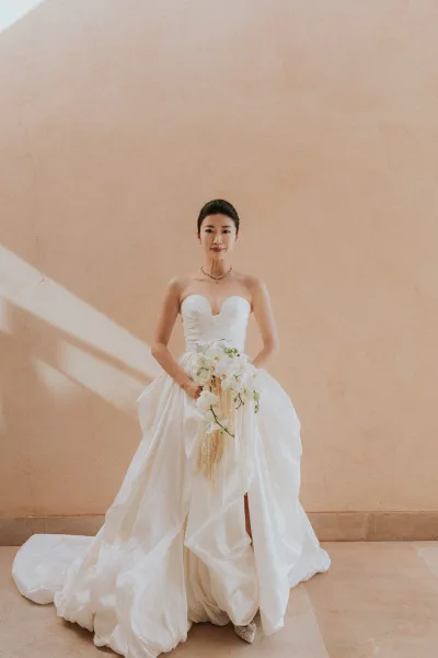 Bridal portrait of a bride in a strapless wedding dress with sweetheart neckline, holding a cascading white orchid bouquet indoors by a neutral wall
