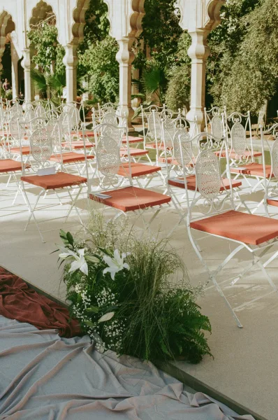 Ceremony seating with outdoor wedding ceremony chairs in white wrought iron, terracotta cushions, and a lily-lined aisle in an arched courtyard colonnade