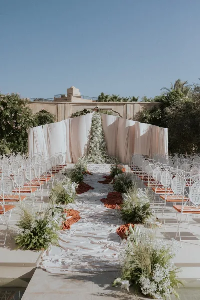 Ceremony setup for an outdoor wedding ceremony with white drapery backdrop, fabric aisle runner, white florals and greenery in a garden courtyard