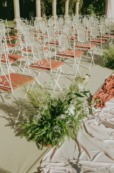 Ceremony seating with outdoor ceremony chairs in white wrought iron, terracotta cushions, and rust fabric aisle with baby’s breath florals under a pergola