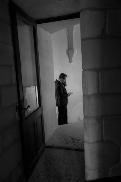 Groom reads a letter in his suit beneath an arched stone ceiling, pausing alone before the ceremony.