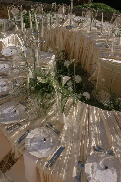 Champagne satin tablecloths and calla lilies adorn reception tables beside a stone wall amid garden greenery