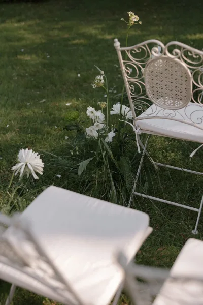 White florals and greenery line the ceremony aisle, with wrought-iron chairs on a garden lawn