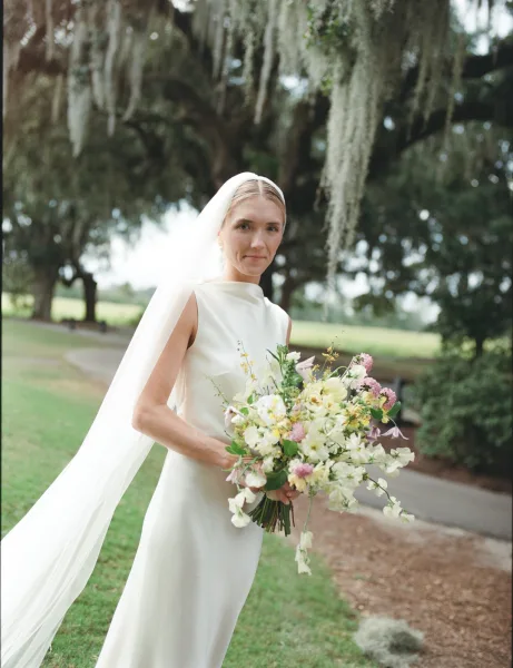 Bridal portrait of a bride holding bouquet in a minimalist high-neck wedding dress, long veil and earrings beneath oak trees with moss