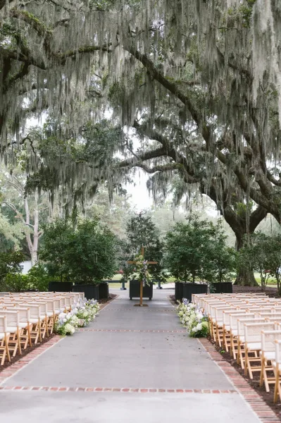 Outdoor ceremony setup with ceremony aisle flowers leading to a wooden cross altar beneath oak trees draped in hanging moss
