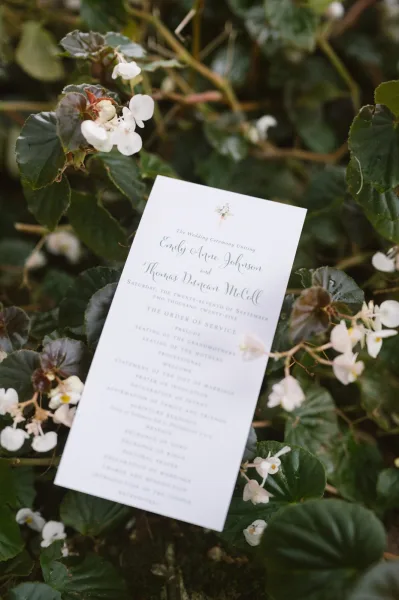 Wedding program card with calligraphy text and floral illustration, tucked among white flowers and greenery against garden foliage background