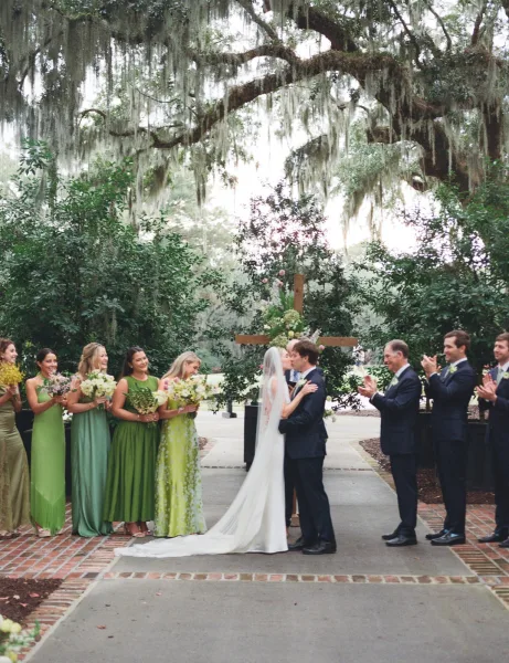 Wedding kiss at the ceremony kiss moment as bride and groom embrace under oak trees with moss, bridal veil and floral cross altar behind