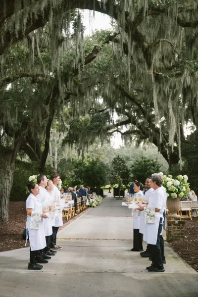 Wedding reception entrance with a champagne welcome line as servers in white aprons offer tray-passed glasses along an oak-lined garden path