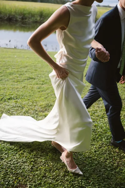 Wedding couple portrait of bride and groom walking hand in hand, bride lifting satin dress train on a grassy lawn by a pond