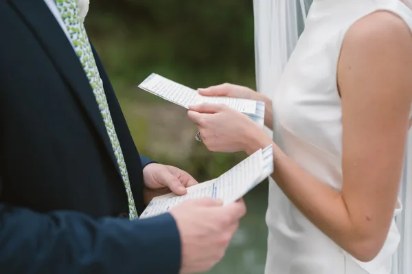 Wedding vows being read from personal vow cards by bride in bridal gown and groom in suit, rings visible at a lakeside ceremony with white drapery