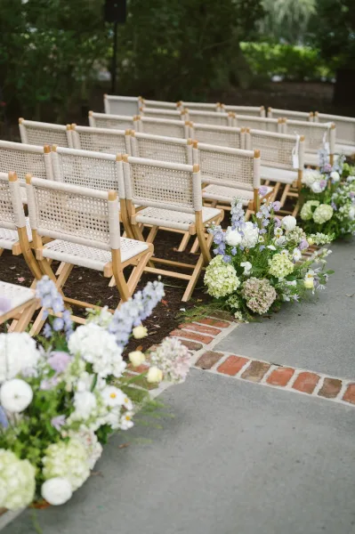 Ceremony aisle decor with floral lined wedding aisle of low hydrangeas, delphinium, and roses beside wicker chairs on a brick-edged path