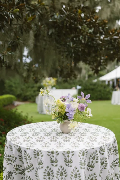 Cocktail table decor with a small floral centerpiece in a ceramic vase on a patterned tablecloth, set on a garden lawn under a white tent