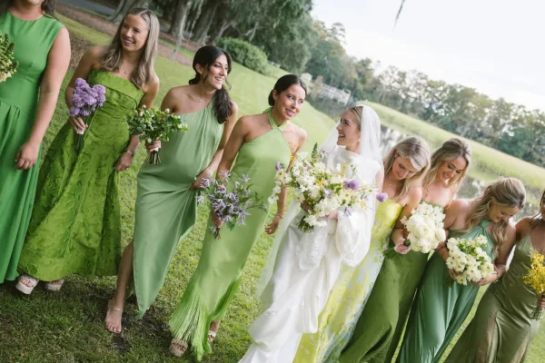 Bride with bridesmaids in a bridal party portrait walking on a grass lawn by a pond, holding wildflower bouquets under a cloudy sky