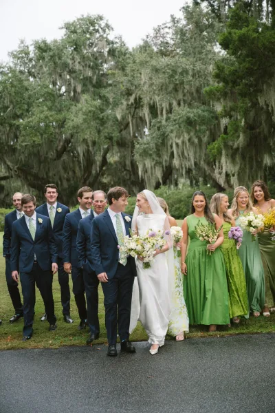 Wedding party portrait of bride and groom walking with bridesmaids in green dresses and groomsmen in navy suits under oak trees with moss