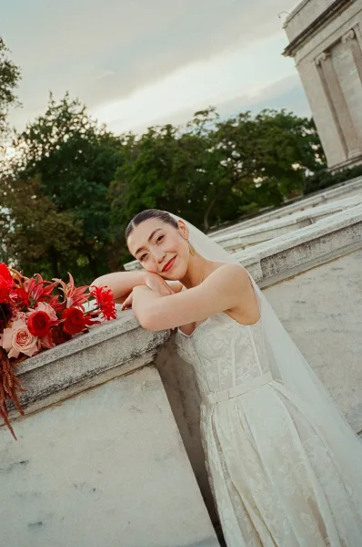 Bridal portrait of bride leaning on railing in a sleeveless textured wedding dress, holding a red and pink rose bouquet on stone steps