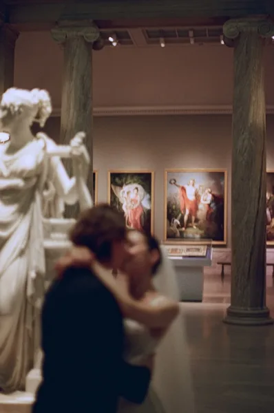 Wedding kiss as bride and groom embrace under a flowing veil, framed by tall columns and statues in a museum gallery hall