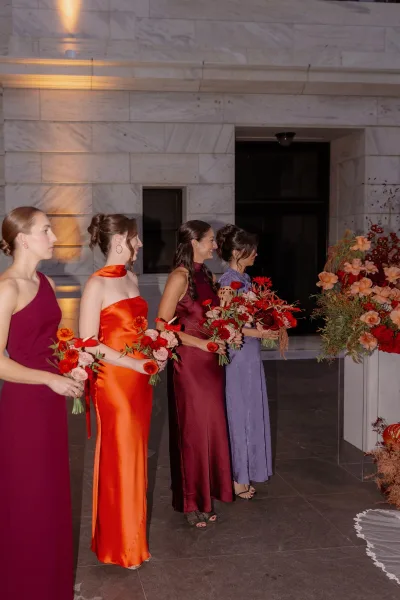 Bridesmaid lineup with bridesmaids holding bouquets of red flowers and pink roses, in mismatched dresses by a marble wall indoors