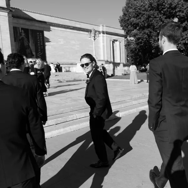 Groom portrait in sunglasses wearing a black tuxedo with bow tie and boutonniere, walking on stone steps by a museum plaza with guests behind