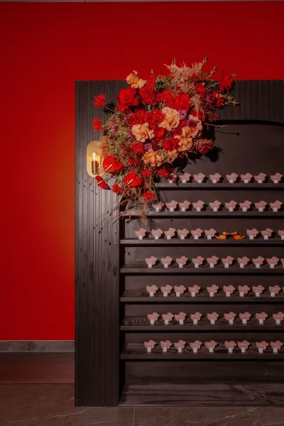 Wedding escort card display with escort card wall shelves, roses and greenery beside a wall sconce against a dramatic red and black backdrop
