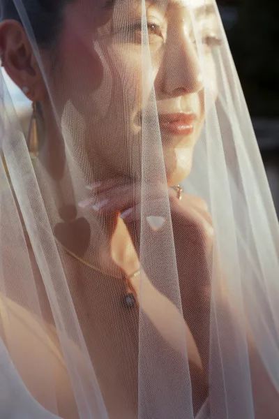 Bridal portrait of a bride under veil, close-up side profile with gold hoop earrings, hand on face, and dark foliage behind