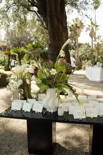 Escort card display with wedding seating cards in classic script beside a calla lily centerpiece on a mirrored table in a garden courtyard