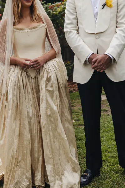 Wedding couple portrait of bride and groom standing hand in hand, her veil and strapless gown beside his white tuxedo, on a garden lawn
