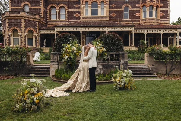 Wedding kiss as bride and groom embrace, her cathedral veil and long train flowing by stone steps of a historic brick estate